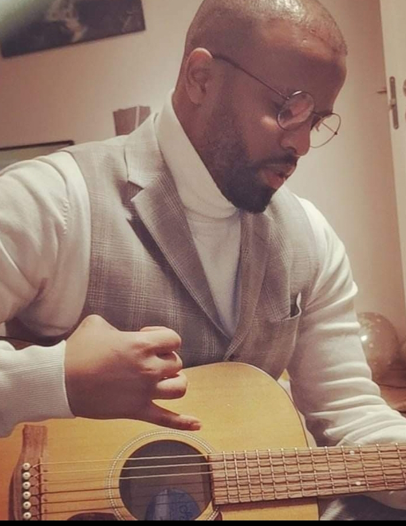 Farid er kledd i vest og skjorte, han klimprer på gitar 