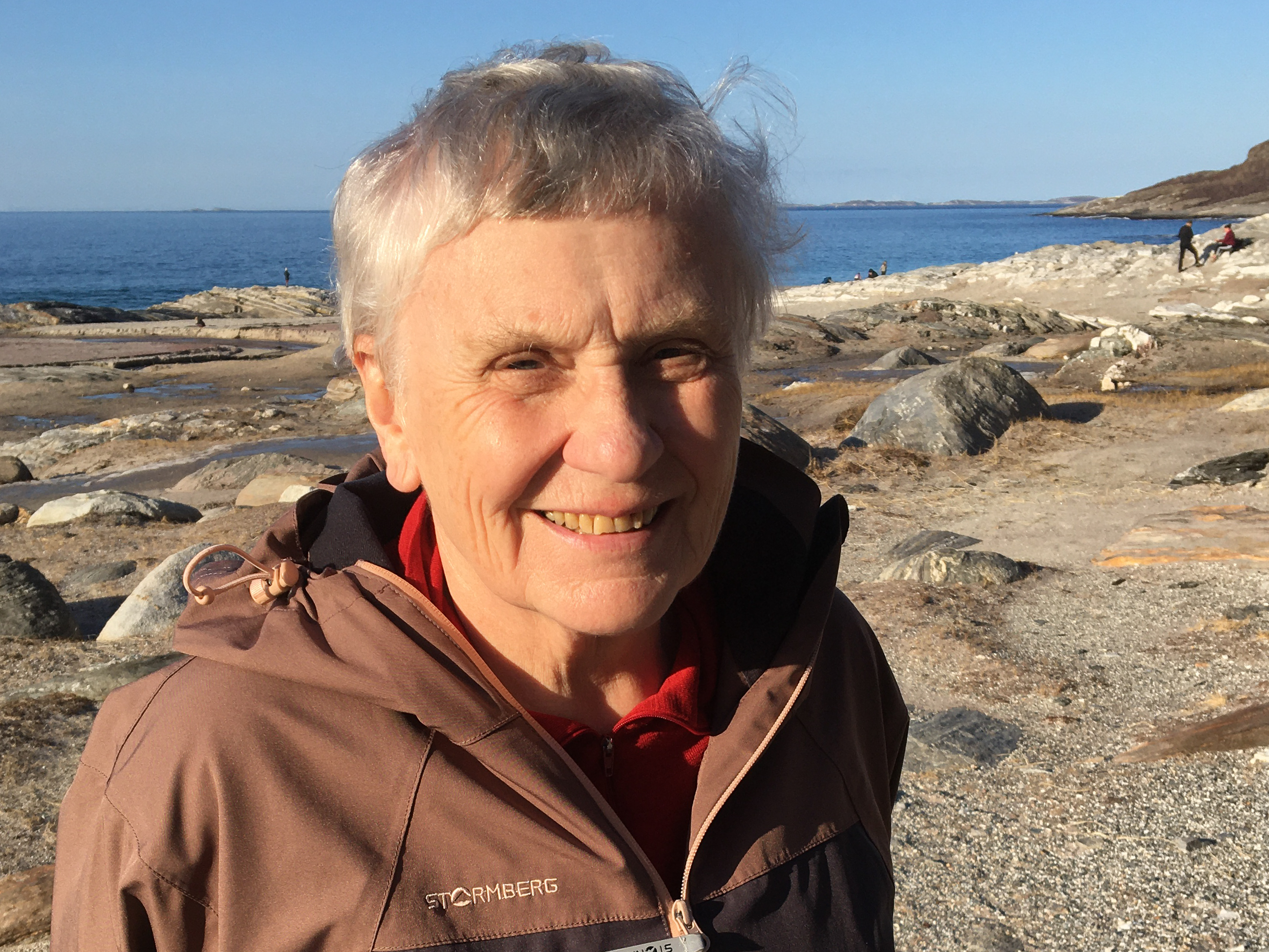 Bilde av eldre kvinne fra skuldre og opp mot grå bakgrunn. Hun har hvit t-skjorte og lyst hår.